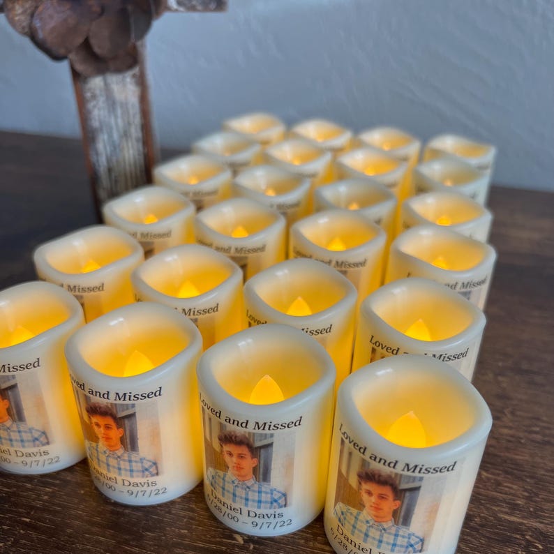 May include: A collection of battery-operated LED candles with a warm, flickering glow. Each candle is white and features a printed photograph and the text "Loved and Missed" along with a name and date. The candles are arranged on a wooden surface.
