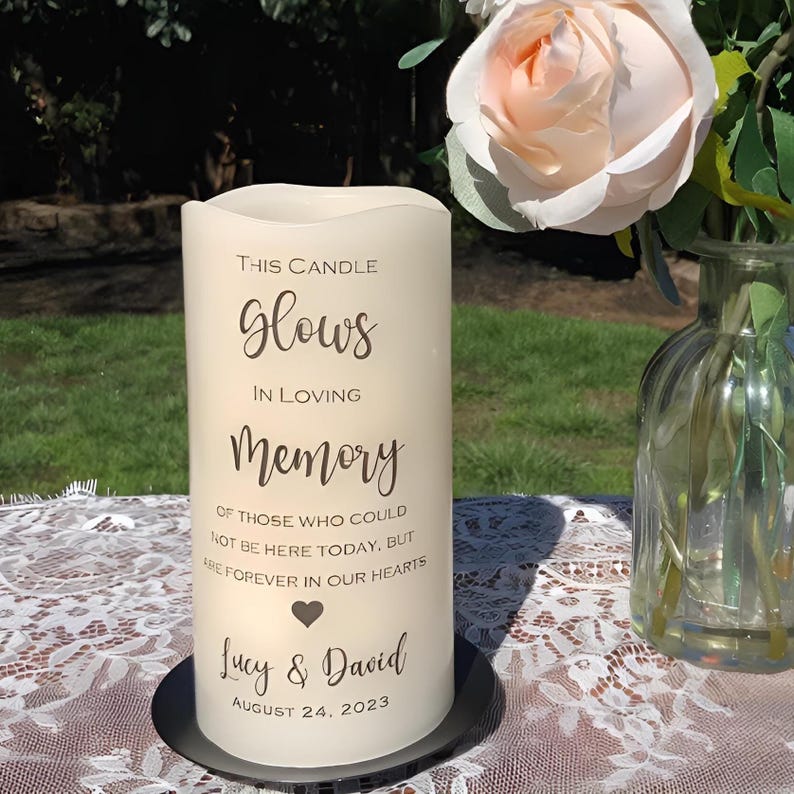 May include: A cream-colored pillar candle with the words "This Candle Glows In Loving Memory" printed in elegant script. The candle includes the names "Lucy & David" and the date August 24, 2023. A black metal base supports the candle.
