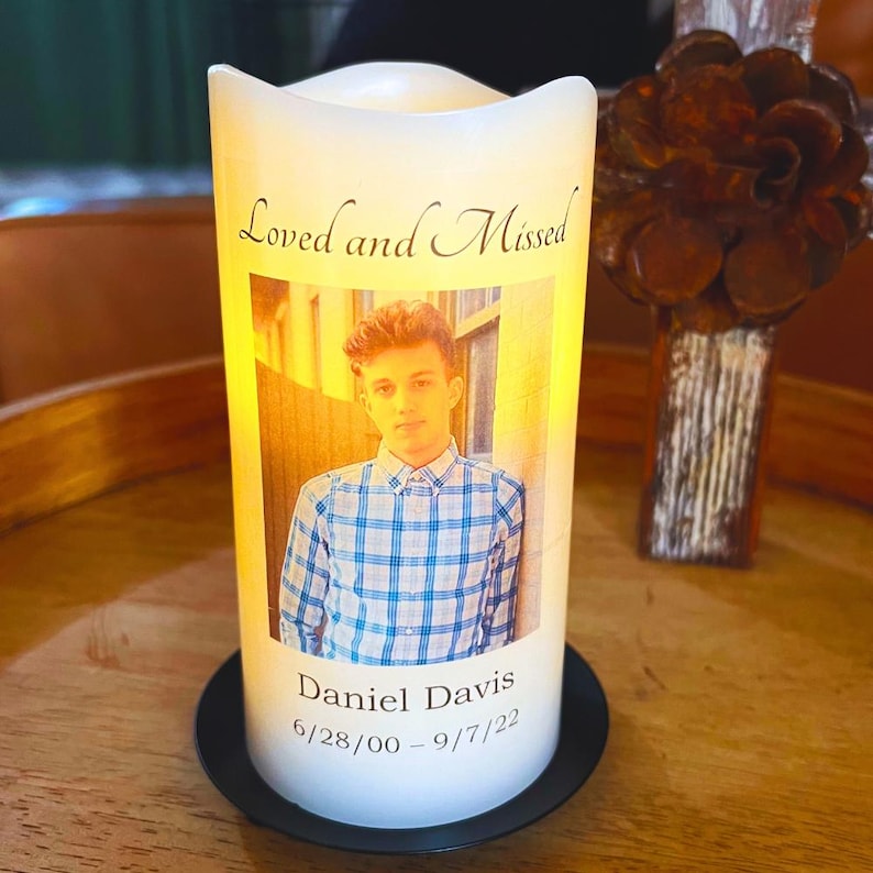 May include: A white LED candle with a photo of a young man in a blue plaid shirt. The candle has the text "Loved and Missed" and "Daniel Davis 6/28/00 - 9/7/22". The candle sits on a black base.