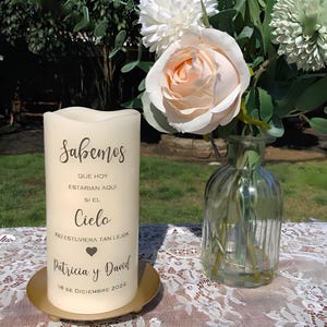 May include: Ivory pillar candle with Spanish script text, on a gold plate. The candle has the text "Sabemos" and other Spanish text. A glass vase with flowers is to the right.