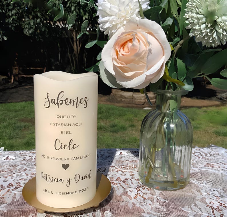 May include: Ivory pillar candle with Spanish script text, on a gold plate. The candle has the text "Sabemos" and other Spanish text. A glass vase with flowers is to the right.