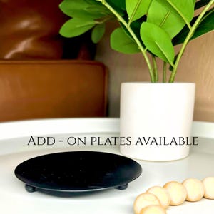 Peut inclure: Une assiette noire de forme ovale avec de petits pieds, pr&eacute;sent&eacute;e sur une surface blanche. Le texte "ADD - ON PLATES AVAILABLE" est visible. Un pot en c&eacute;ramique blanc avec une plante verte est en arri&egrave;re-plan.
