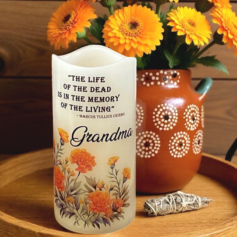 K&ouml;nnte beinhalten: Eine wei&szlig;e Kerze mit dem Text "THE LIFE OF THE DEAD IS IN THE MEMORY OF THE LIVING" und dem Wort "Grandma". Die Kerze ist mit orangefarbenen Blumenmustern verziert. Eine Vase mit orangefarbenen Blumen und ein Salbeib&uuml;ndel sind ebenfalls zu sehen.