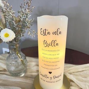 May include: A white LED candle with the text "Esta vela Brilla" and a dedication to Camilla and Daniel, dated August 26, 2026. The candle sits on a gold-coloured base, with a vase of flowers in the background.