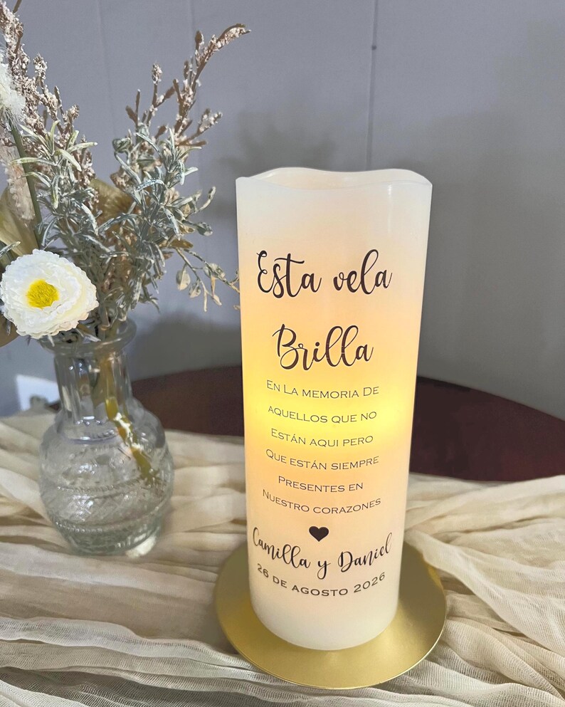 May include: A white LED candle with the text "Esta vela Brilla" and a dedication to Camilla and Daniel, dated August 26, 2026. The candle sits on a gold-coloured base, with a vase of flowers in the background.