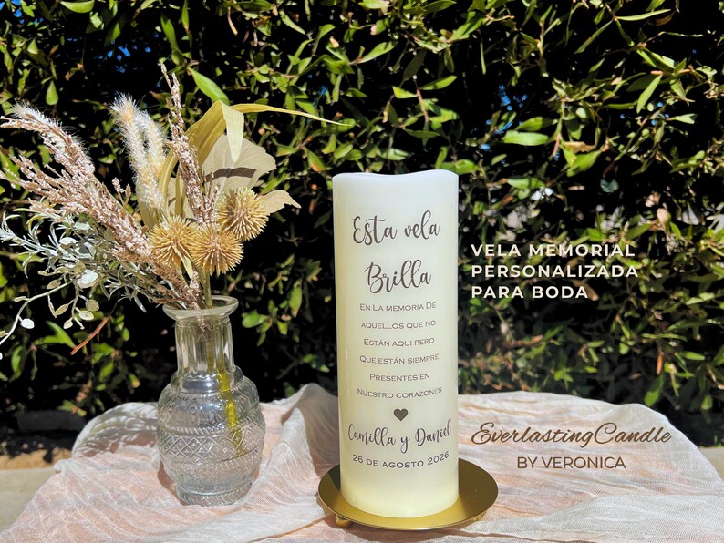 May include: A white pillar candle with text, on a gold-coloured base. A small glass vase with dried flowers is next to the candle. The text on the candle reads "Esta vela Brilla" and "Vela Memorial Personalizada Para Boda."