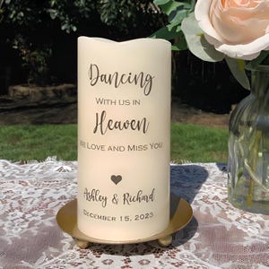 Peut inclure: Une bougie pilier cr&egrave;me avec l'inscription "Dancing With Us In Heaven, We Love and Miss You." En dessous, un c&oelig;ur, les noms "Ashley & Richard" et la date "December 15, 2023." La bougie repose sur un socle en m&eacute;tal dor&eacute;.
