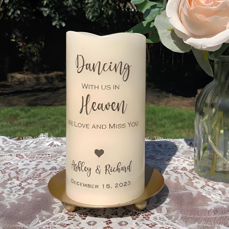 Peut inclure: Une bougie pilier cr&egrave;me avec l'inscription "Dancing With Us In Heaven, We Love and Miss You." En dessous, un c&oelig;ur, les noms "Ashley & Richard" et la date "December 15, 2023." La bougie repose sur un socle en m&eacute;tal dor&eacute;.