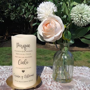 May include: A cream candle with Spanish text, including the word "Porque", names, and a date. The candle is on a gold-coloured metal plate. A bouquet of flowers and a glass vase are in the background, on a lace tablecloth.