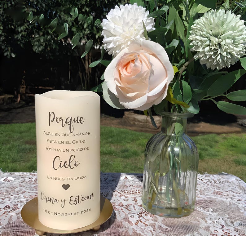 May include: A cream candle with Spanish text, including the word "Porque", names, and a date. The candle is on a gold-coloured metal plate. A bouquet of flowers and a glass vase are in the background, on a lace tablecloth.