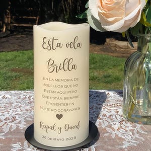 Peut inclure: Une bougie pilier cr&egrave;me avec l'inscription "Esta vela Brilla" et un message comm&eacute;moratif en espagnol. La bougie repose sur une base en m&eacute;tal noir, avec un arrangement floral en arri&egrave;re-plan. La date "26 DE MAYO 2023" est &eacute;galement imprim&eacute;e sur la bougie.