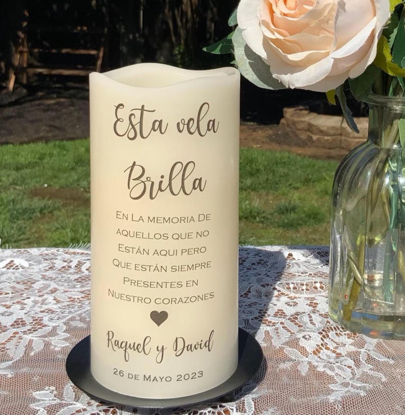 Peut inclure: Une bougie pilier cr&egrave;me avec l'inscription "Esta vela Brilla" et un message comm&eacute;moratif en espagnol. La bougie repose sur une base en m&eacute;tal noir, avec un arrangement floral en arri&egrave;re-plan. La date "26 DE MAYO 2023" est &eacute;galement imprim&eacute;e sur la bougie.