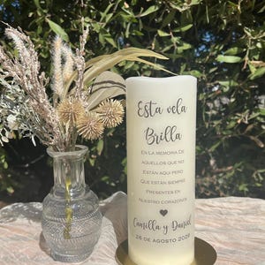 May include: A white pillar candle with Spanish text, including "Esta vela Brilla". The candle is on a gold-coloured base. A small glass vase with dried flowers is to the left. The date 26 de Agosto 2026 is visible.