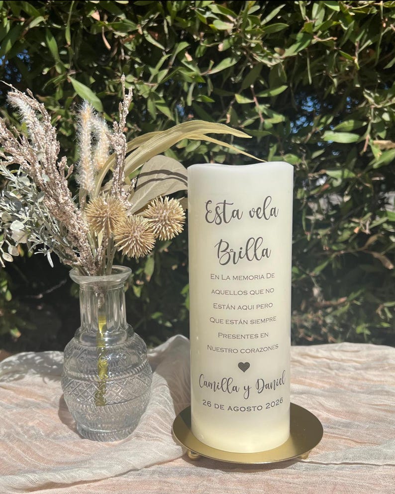 May include: A white pillar candle with Spanish text, including "Esta vela Brilla". The candle is on a gold-coloured base. A small glass vase with dried flowers is to the left. The date 26 de Agosto 2026 is visible.