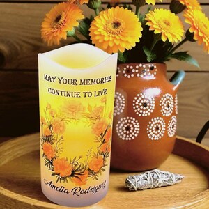 K&ouml;nnte beinhalten: Eine dekorative Kerze mit dem Text "MAY YOUR MEMORIES CONTINUE TO LIVE" und dem Namen "Amelia Rodriguez". Die Kerze hat ein Blumendesign und steht neben einer Vase mit gelben Blumen.