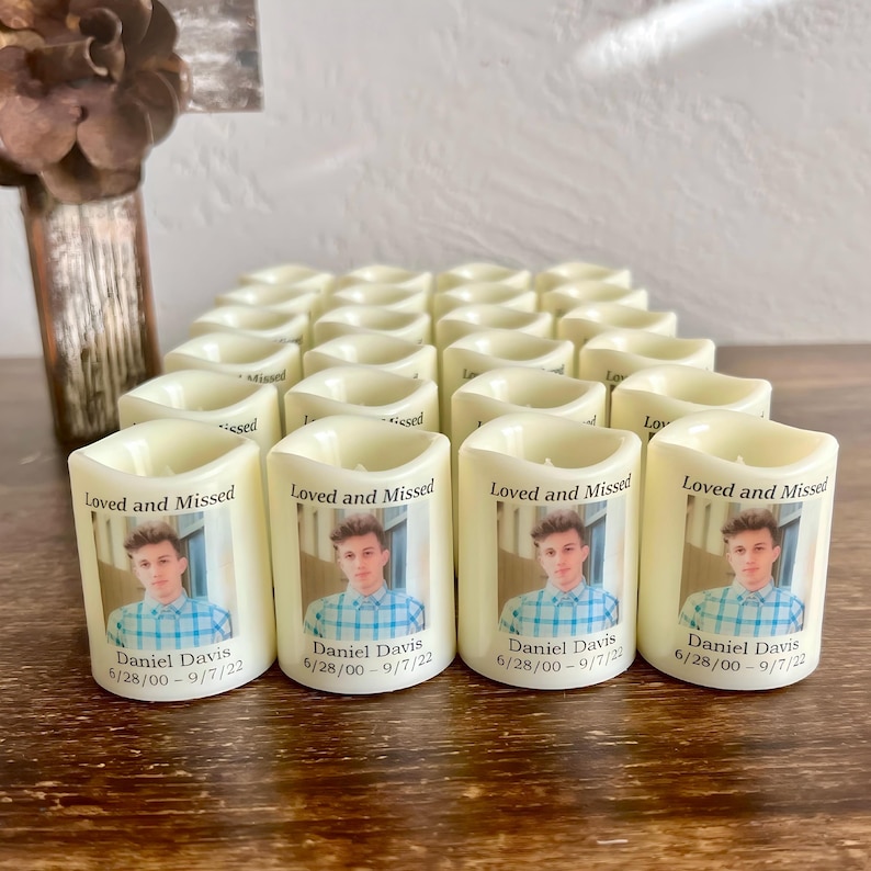 Op de afbeelding: Een verzameling gebroken witte, cilindervormige LED-herdenkingskaarsen met een afgedrukte foto van een jong persoon in een blauw geruit overhemd. Elke kaars toont de tekst Loved and Missed, de naam Daniel Davis en de data 6/28/00 - 9/7/22.