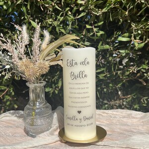 May include: A cream-coloured pillar candle with Spanish text, including "Esta vela Brilla". The candle is on a gold-coloured metal plate. A small vase with dried flowers is to the left. The date 26 de agosto 2026 is visible.