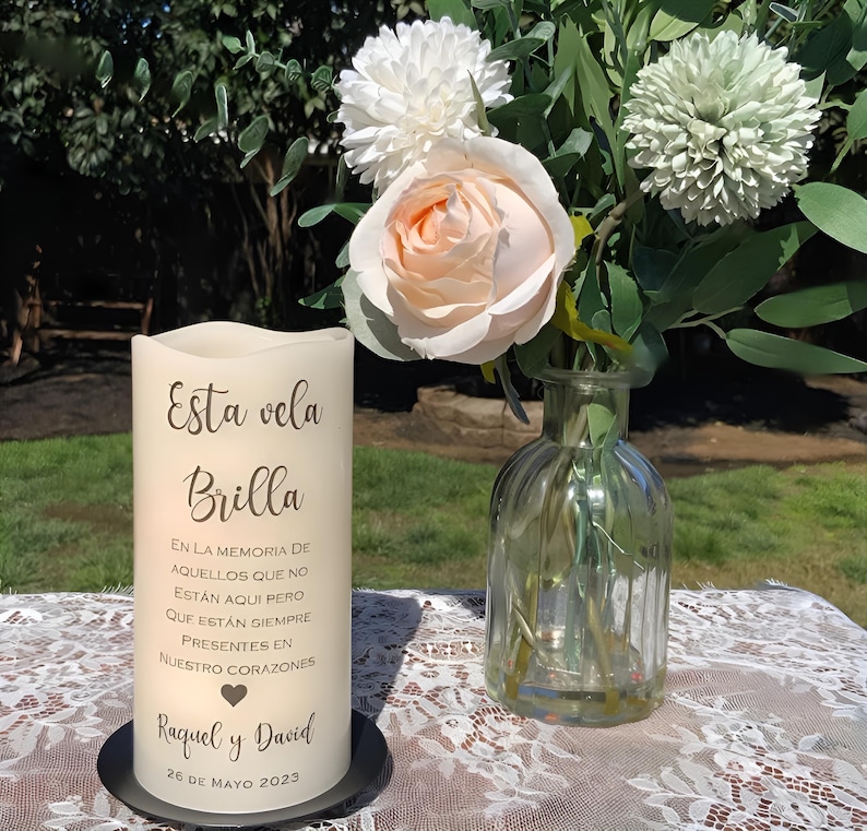 May include: A cream-colored pillar candle with the text "Esta vela Brilla" and other Spanish text. The candle sits on a black base, next to a glass vase with flowers. A bouquet of white and peach flowers is in the vase.