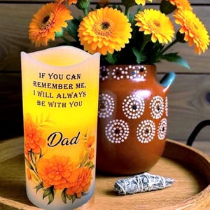 K&ouml;nnte beinhalten: Eine dekorative Kerze mit dem Text "IF YOU CAN REMEMBER ME, I WILL ALWAYS BE WITH YOU Dad" und floralen Akzenten. Neben der Kerze steht eine braune Vase mit wei&szlig;en Punkten und gelben Blumen. Ein Holztablett h&auml;lt die Gegenst&auml;nde.