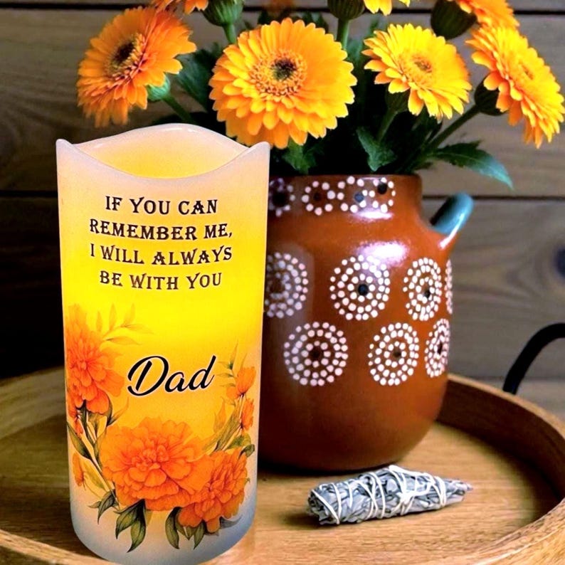 K&ouml;nnte beinhalten: Eine dekorative Kerze mit dem Text "IF YOU CAN REMEMBER ME, I WILL ALWAYS BE WITH YOU Dad" und floralen Akzenten. Neben der Kerze steht eine braune Vase mit wei&szlig;en Punkten und gelben Blumen. Ein Holztablett h&auml;lt die Gegenst&auml;nde.