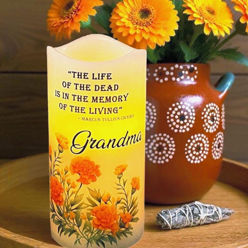 May include: A yellow candle with the quote "THE LIFE OF THE DEAD IS IN THE MEMORY OF THE LIVING" and the word "Grandma". The candle has orange floral accents and is next to a vase of orange flowers.