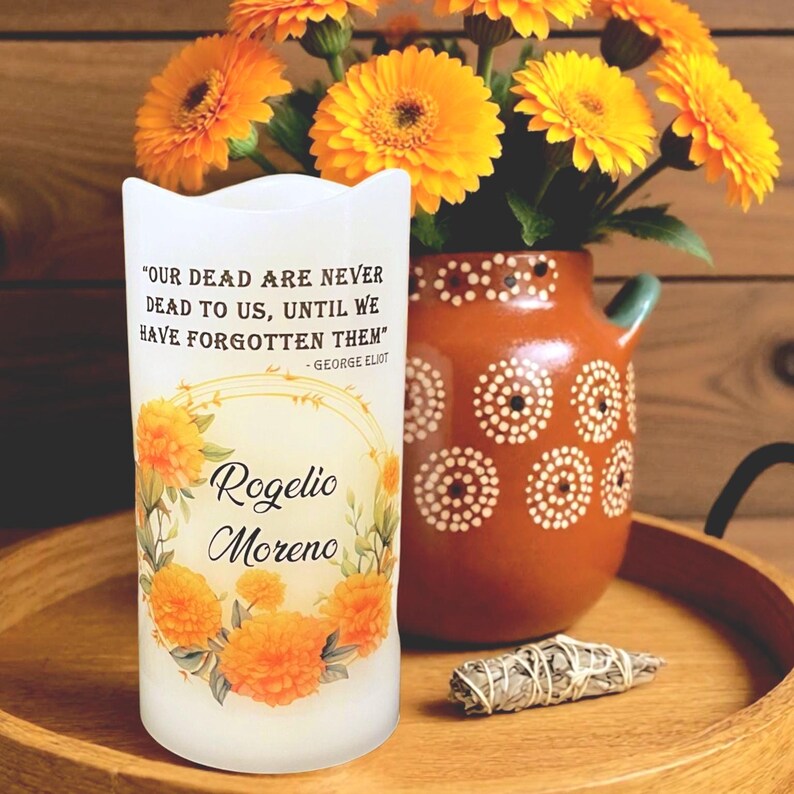 K&ouml;nnte beinhalten: Eine wei&szlig;e Kerze mit dem Zitat "OUR DEAD ARE NEVER DEAD TO US, UNTIL WE HAVE FORGOTTEN THEM" und dem Namen "Rogelio Moreno". Die Kerze ist mit orangefarbenen Blumen verziert und steht auf einem Holztablett. Im Hintergrund steht eine Vase mit orangefarbenen Blumen.