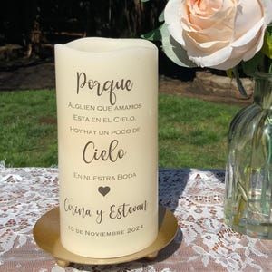 Peut inclure: Une bougie pilier cr&egrave;me avec du texte en espagnol, dont "Porque", "Cielo", et les noms "Corina y Estevan" avec une date. La bougie est pos&eacute;e sur une plaque m&eacute;tallique dor&eacute;e. Un arrangement floral est en arri&egrave;re-plan.
