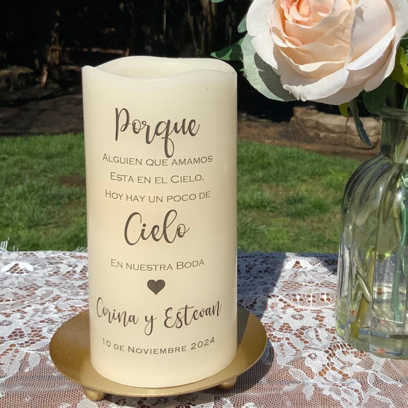 Peut inclure: Une bougie pilier cr&egrave;me avec du texte en espagnol, dont "Porque", "Cielo", et les noms "Corina y Estevan" avec une date. La bougie est pos&eacute;e sur une plaque m&eacute;tallique dor&eacute;e. Un arrangement floral est en arri&egrave;re-plan.
