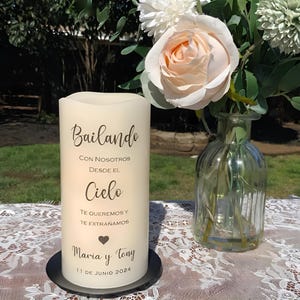 May include: A cream-coloured pillar candle with Spanish text, including "Bailando". The candle is on a black base, next to a vase of white and peach flowers. The date "11 DE JUNIO 2024" is visible.