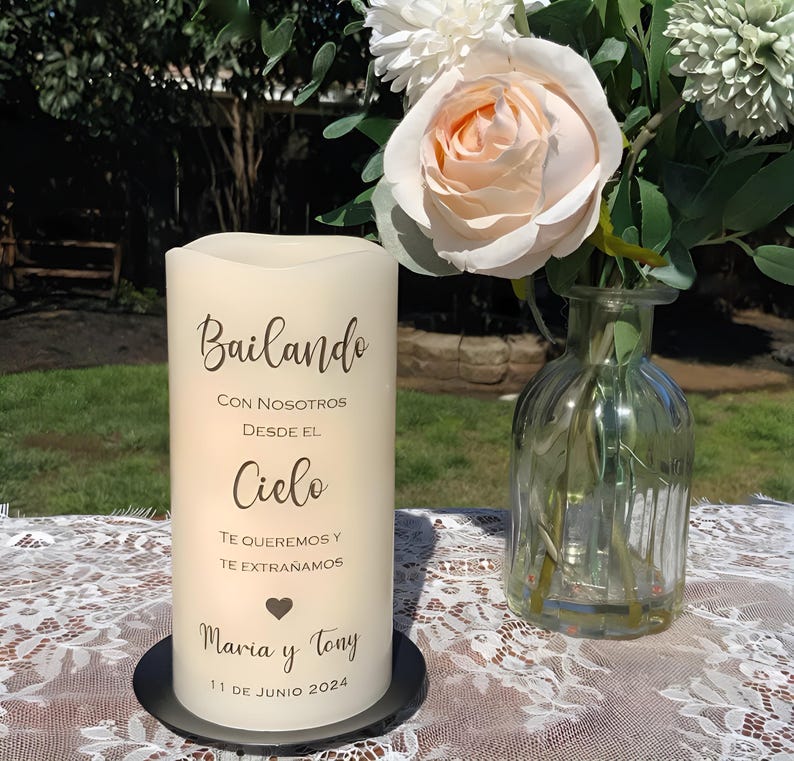 May include: A cream-coloured pillar candle with Spanish text, including "Bailando". The candle is on a black base, next to a vase of white and peach flowers. The date "11 DE JUNIO 2024" is visible.