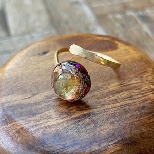 May include: A gold-toned adjustable ring featuring a large, round, iridescent stone. The stone displays a mix of colors, including purple, gold, and red. The ring's band has a hammered texture.