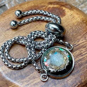 May include: A silver-toned adjustable bracelet featuring a round pendant with a dark, textured interior containing gold-colored metallic shavings, white granular material, and small iridescent flakes that catch the light.