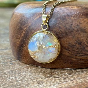 May include: A round pendant necklace featuring a gold-toned metal setting and a chain. The circular pendant contains a mosaic of iridescent white and blue stone fragments interspersed with thin, reflective gold-colored metallic flakes.