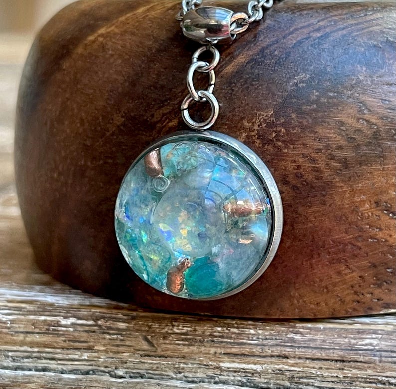 May include: A round pendant necklace with a silver-colored chain. The pendant has a clear, domed glass front, revealing a mix of blue, green, and copper-colored elements. The pendant is set in a silver-colored metal frame.