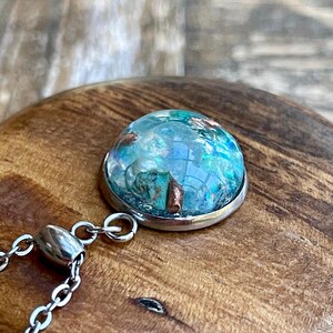 May include: A round pendant with a silver-colored metal rim and a clear, domed center. The center features a mix of blue, green, and copper-colored elements, creating a unique, textured appearance. A silver chain is attached to the pendant.