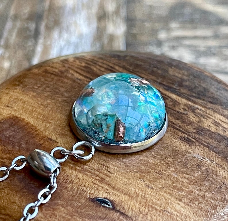 May include: A round pendant with a silver-colored metal rim and a clear, domed center. The center features a mix of blue, green, and copper-colored elements, creating a unique, textured appearance. A silver chain is attached to the pendant.