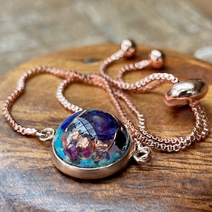 May include: A rose gold bracelet with a round, clear pendant. The pendant contains a mix of blue, purple, and copper-colored materials. The bracelet is on a wooden surface.