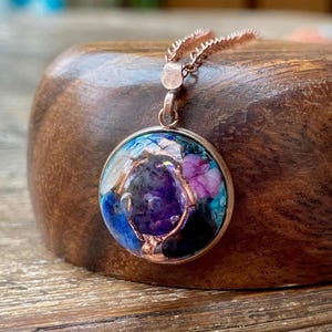 May include: A round pendant necklace with a copper-colored chain and setting. The pendant features a mix of colors, including purple, blue, pink, and black, with a shiny, reflective surface. The necklace is displayed on a wooden surface.