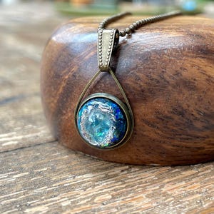 May include: A pendant necklace featuring a circular bronze-toned setting with a textured, metallic rim. The center contains a vibrant, iridescent blue and teal stone-like inlay with shimmering silver-toned flecks, suspended from a beaded chain.