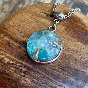 May include: A round pendant necklace with a silver chain. The pendant features a clear, circular resin design with blue and iridescent elements, and copper accents. The necklace is displayed on a wooden surface.