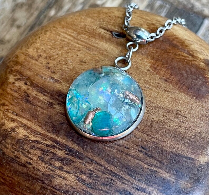 May include: A round pendant necklace with a silver chain. The pendant features a clear, circular resin design with blue and iridescent elements, and copper accents. The necklace is displayed on a wooden surface.