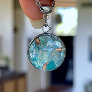 May include: A round pendant necklace with a silver chain. The pendant has a clear, circular glass or resin casing filled with blue and teal-colored elements and copper-colored accents. The necklace is held up by a finger.