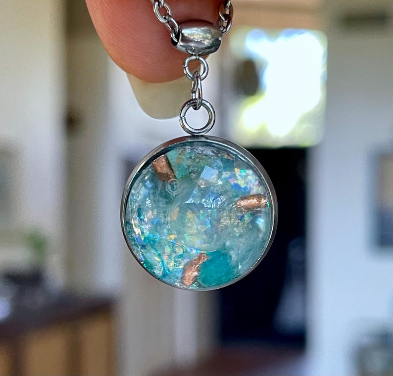 May include: A round pendant necklace with a silver chain. The pendant has a clear, circular glass or resin casing filled with blue and teal-colored elements and copper-colored accents. The necklace is held up by a finger.
