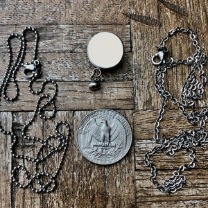May include: A silver-toned necklace with a ball chain and a lobster clasp, next to a round pendant with a blank white surface. A quarter coin is also present, showing the eagle design.