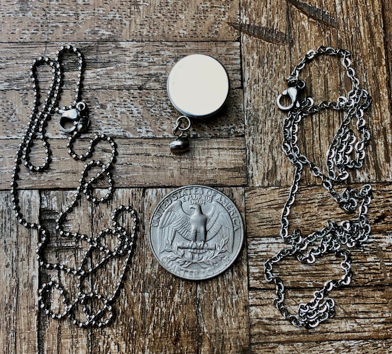 May include: A silver-toned necklace with a ball chain and a lobster clasp, next to a round pendant with a blank white surface. A quarter coin is also present, showing the eagle design.