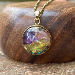 May include: A round gold-tone pendant necklace featuring a resin-filled center with embedded purple, pink, and yellow stone fragments accented by shimmering gold leaf flakes. The pendant hangs from a matching gold-tone chain link necklace.