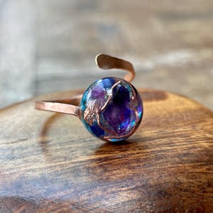 May include: A copper-toned ring with a large, round, iridescent stone. The stone features shades of blue, purple, and hints of gold. The ring's band is a simple, open design, with a curved accent above the stone.