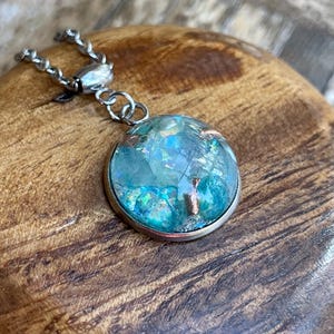 May include: A round pendant necklace with a silver chain. The pendant has a clear, domed glass encasing a blue and green iridescent design with copper accents. The pendant is set in a silver bezel.