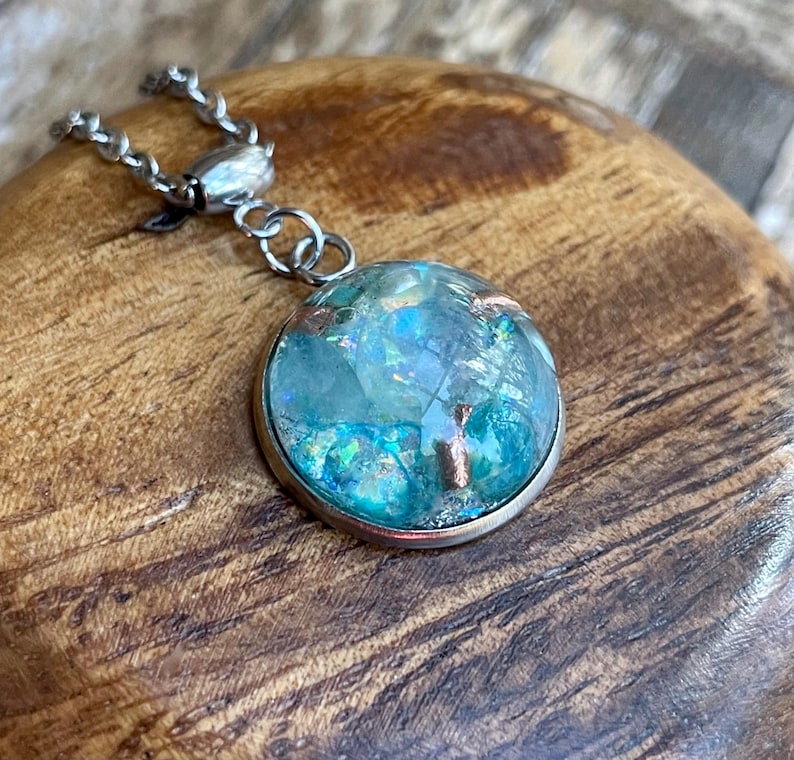 May include: A round pendant necklace with a silver chain. The pendant has a clear, domed glass encasing a blue and green iridescent design with copper accents. The pendant is set in a silver bezel.