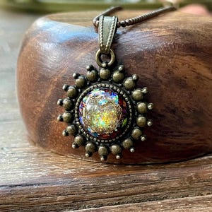 May include: A round pendant necklace featuring a central iridescent, multi-colored stone set in a dark bronze-toned metal frame. The frame is decorated with a series of small, raised spheres arranged in a sunburst pattern around the center.